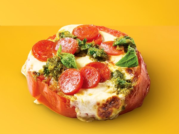 Melted cheese, mini pepperoni, and basil pesto topped on a slice of tomato on a yellow background. 