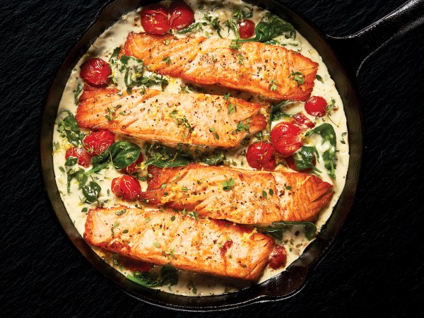 Four salmon fillets in a cast-iron skillet of creamy sauce, tomatoes, and spinach on a black background. 