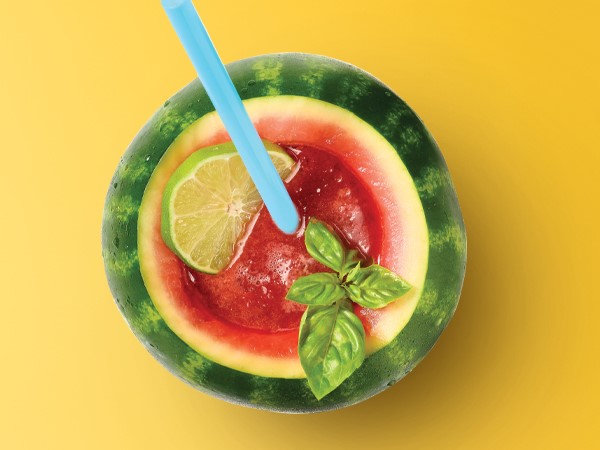 Watermelon beverage in a hollowed out mini watermelon with a lime slice and blue straw on a yellow background. 