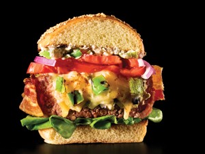 Tall burger cut in half showing melted cheese, burger, lettuce, tomato and onion slices in between a sesame seed bun on a black background.