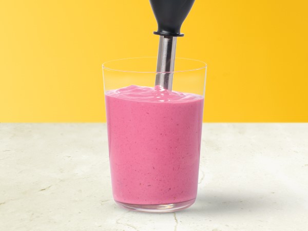 A pink-purple smoothie in a glass with an immersion blender inserted in smoothie on a yellow and white background.