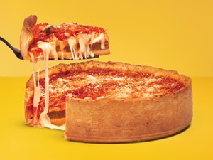 Chicago-style pizza with one slice raised on a black spatula above the pizza on a yellow background. 