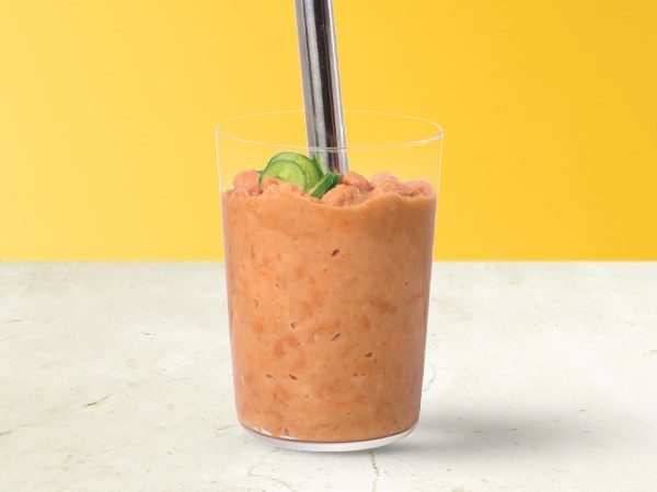 Light-orange colored chili-refried beans in a glass with an immersion blender inserted on a white and yellow background.