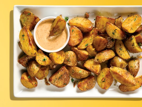 A white serving plate with roasted potato wedges next to a ramekin of dipping sauce on a yellow background. 
