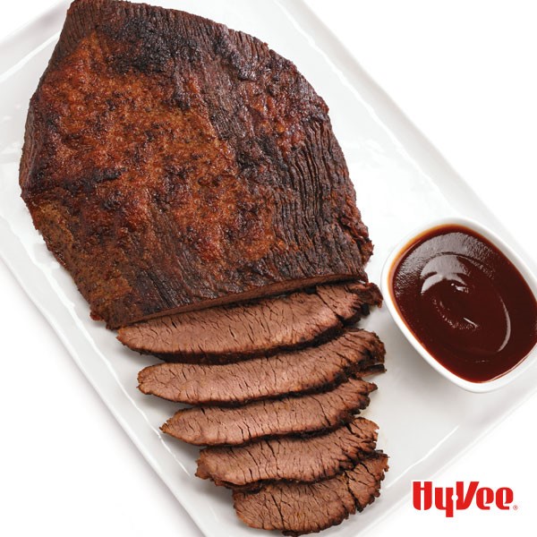 Platter of a partially-sliced beef brisket with a side dish of barbecue sauce