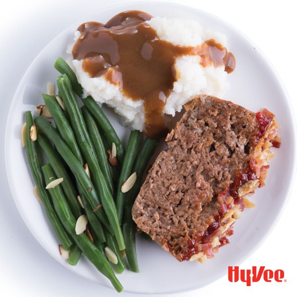 White plate with green beans sprinkled with almonds, mashed potatoes with gravy, and meatloaf slice