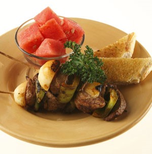 Steak kabobs on a plate with toast and a bowl of watermelon
