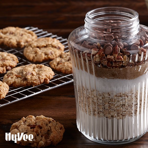 Jar filled with four, sugar, oats, pecan pieces, and chocolate chips next to baked cookies on wire rack