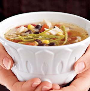 Hands holding white bowl of soup filled with chicken, black beans, corn, and thinly sliced tortilla strips