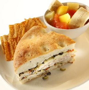 Muffaletta sandwich with olives, chicken, and focaccia bread with a side of mixed fruit and chips