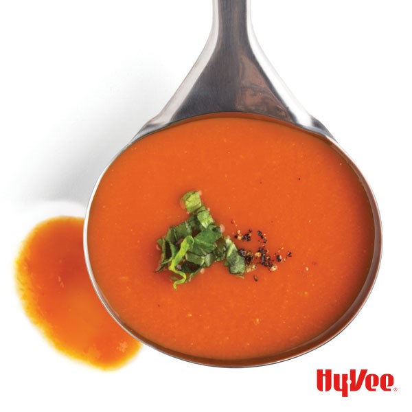 Ladle holding tomato soup with minced basil and black pepper for garnish