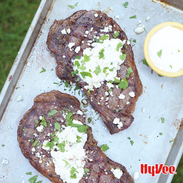 Tray of rib eye steaks topped with blue cheese crumbles and cilantro with a side of horseradish sauce