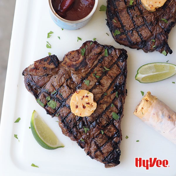 Grilled porterhouse steak  topped with chipotle butter, sprinkled with parsley on a white plate with sliced limes