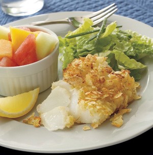 Potato chip crusted cod on a plate with chopped mixed fruit, lemon wedge, and lettuce salad
