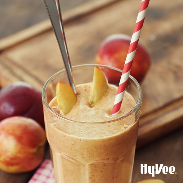 Glass of peach smoothie garnished with peach slices, spoon and red-and-white straw