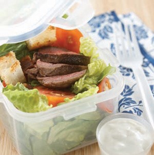 Plastic container filled with salad topped with blue cheese and steak