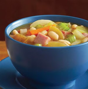 Blue bowl filled with yellow potatoes, white beans, chopped celery, chopped carrots, and diced ham