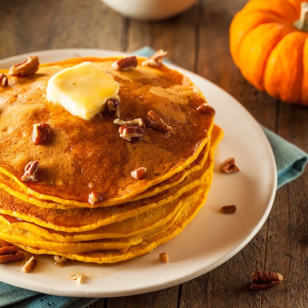 Stack of pumpkin pancakes topped with maple syrup, butter and pecans on a white plate