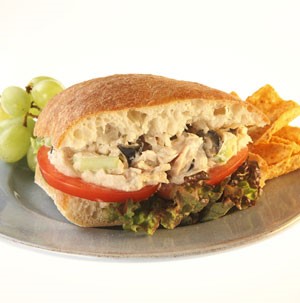Sandwich filled with lettuce, sliced tomato, and tuna salad on a grey plate with chips and green grapes
