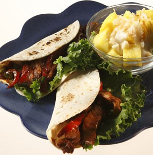 Two flour tortillas filled with sliced pork, peppers, and lettuce, with a side of diced pineapple on a blue plate
