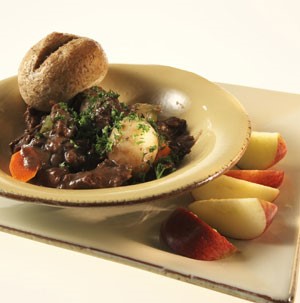 Potato beef stew in a bowl with a dinner roll and a side of sliced apples