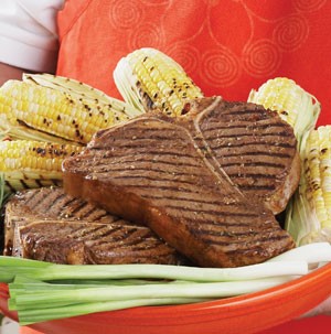 Orange plate held by a man with an orange apron filled with grilled t-bone steaks, grilled corn on the cob and green onions