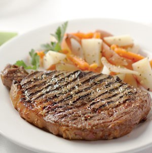 Grilled rib eye steak on a white plate with chopped potatoes, carrots, and fresh parsley