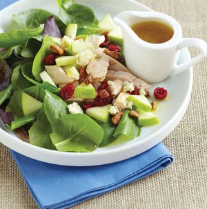 White plate with mixed greens, chopped chicken, diced apples, dried cranberries and dressing on the side