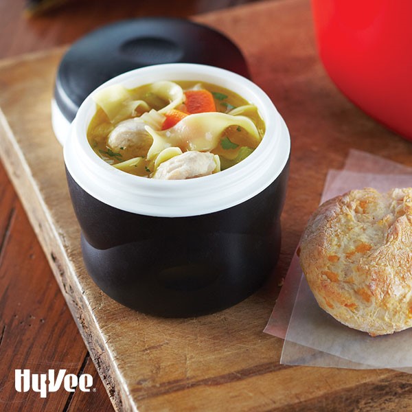 Thermos filled with noodles, parsley, sliced carrots, and chunks of cooked turkey on a wooden cutting board