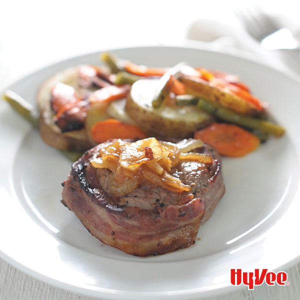 Bacon-wrapped sirloin steak topped with caramelized onions with assorted vegetables on a white plate