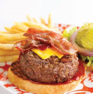 Grilled hamburger bun topped with hamburger patty, cheese, and bacon strips on top