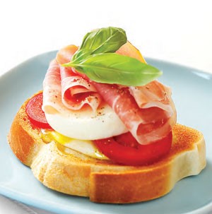 Slice of french bread topped with tomato, mozzarella, prosciutto, egg and basil