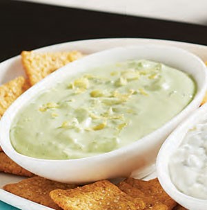 Light green goddess dip in white bowl topped with lemon zest with whole grain chips on the side
