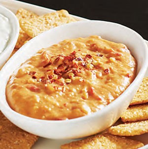 Serving bowl filled with red pepper hummus, topped with crushed red pepper flakes and surrounded by crackers
