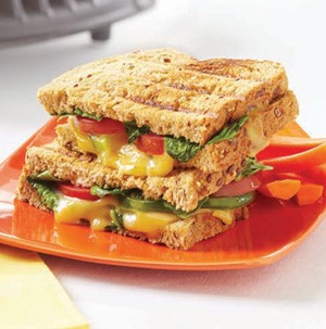 Orange plate topped with grilled bread, melted yellow cheese, spinach, sliced tomatoes, and side of carrot sticks