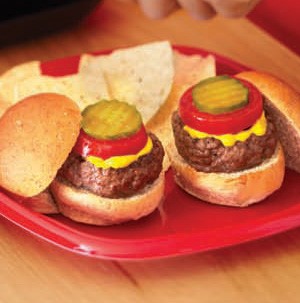 Mini hamburger buns topped with burger patty, yellow mustard, red tomato slice, and pickle slice on a red plate with tortilla chips on the side