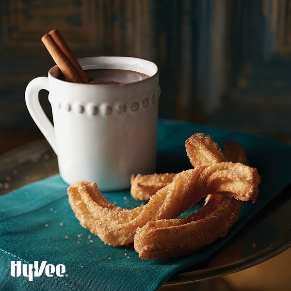 White mug with drinking chocolate and cinnamon stick with churros on the side
