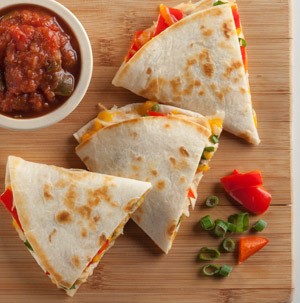 Quesadilla wedges on a wooden cutting board with a side of chopped tomatoes, green onions and a bowl of salsa