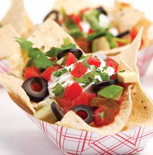 Red and white paper basket filled with tortilla chips, cilantro, sliced black olives, and diced tomatoes