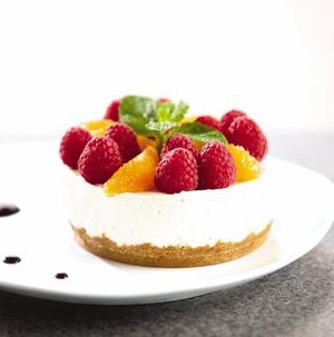 White plate topped with circular tart with graham cracker crust, cream cheese filling and topped with orange segments, whole raspberries and fresh mint