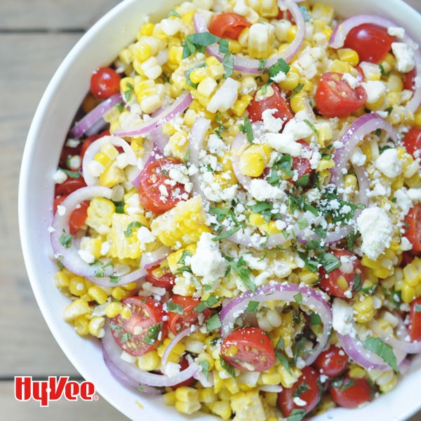 White bowl filled with kernel corn, halved cherry tomatoes, fresh basil, sliced red onions, and crumbled cheese