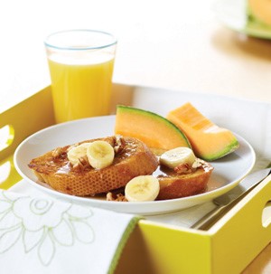 French toast topped with nuts and banana slices on a yellow serving tray with cantaloupe and orange juice