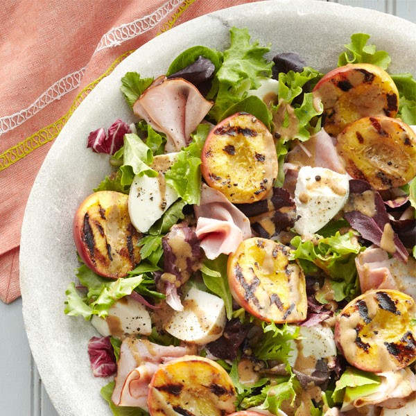 White bowl filled with fresh mozzarella cheese, grilled peach halves, sliced deli ham and mixed greens