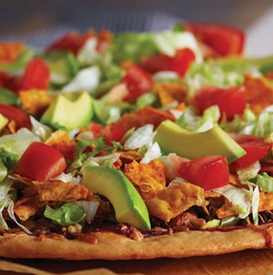 Thin cursted pizza topped with sliced avocado, diced  tomatoes, cooked ground meat, and chips