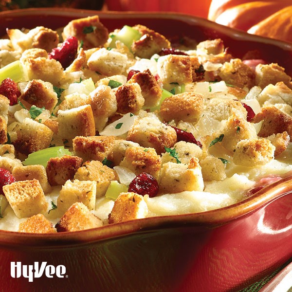 Red circular casserole dish filled with bread cubes, chopped celery, dried cranberries, and chopped onions