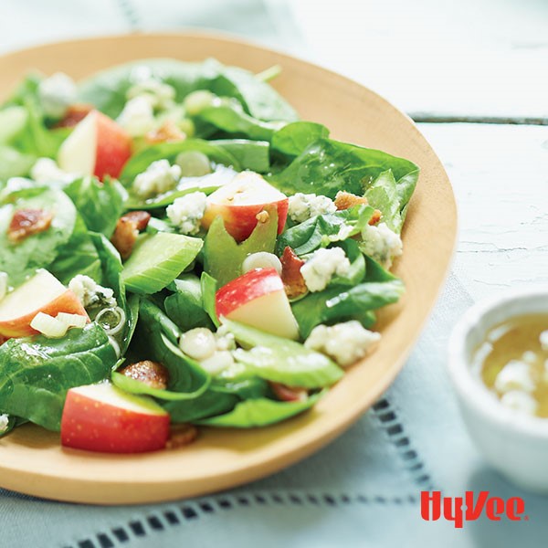 Wooden bowl filled with blue cheese, bacon and apple salad with side of green onion vinaigrette