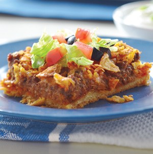 Slice of taco casserole with ground meat, dough crust, and topped with diced tomatoes, sliced black olives, and shredded lettuce