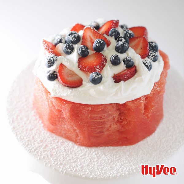 Thick circular piece of watermelon with whipped topping, halved strawberries, whole blueberries, and sprinkled with powdered sugar