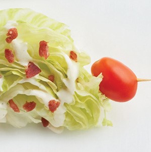 Romaine wedge salad drizzled with white dressing, bacon bits, and a cherry tomato on a skewer