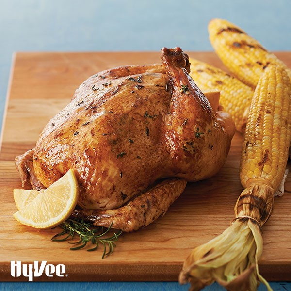 Whole chicken on a wooden cutting board with grilled corn, fresh rosemary, and lemon wedges